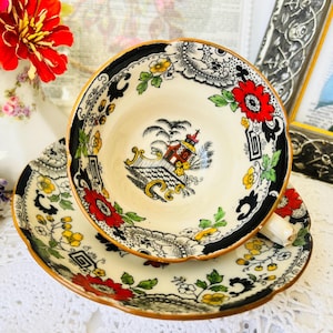 Puede incluir: Un juego de taza y platillo de té vintage. La taza y el platillo de color crema presentan un borde negro y patrones florales con acentos rojos, amarillos y verdes. El interior de la taza muestra una ilustración escénica.