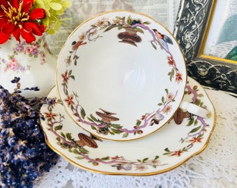 Könnte beinhalten: Weißes Tee-Set mit goldenem Rand. Das Set besteht aus einer Tasse und Untertasse mit floralen und Vogelmotiven in Braun, Grün, Rosa und Rot. Die Tasse steht auf der Untertasse. Wahrscheinlich Vintage.