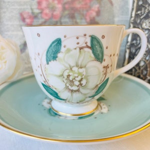 May include: A white teacup and saucer with a floral design. The cup has a green and white floral pattern with a gold rim. The saucer is a light blue color with a gold rim.
