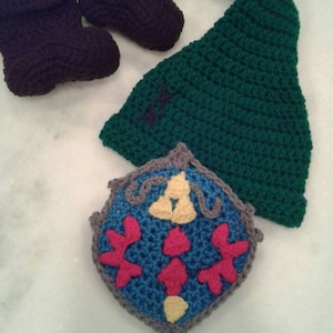 Puede incluir: Sombrero, botines y escudo de crochet para bebé. El sombrero es verde, los botines son negros y el escudo es azul, amarillo y rosa.