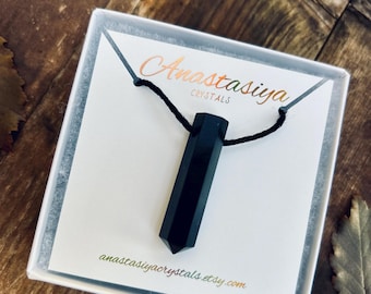 May include: A black crystal pendant necklace displayed in a white box. The pendant is a long, faceted shape suspended on a black cord. The box has the text "Anastasiya Crystals" and the Etsy shop URL printed on it.