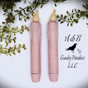 May include: Two pink glitter LED candles with a warm white glow. The candles are approximately 15 cm tall and have a realistic wax look. A&B Country Primitives L.L.C. is printed on the white background.