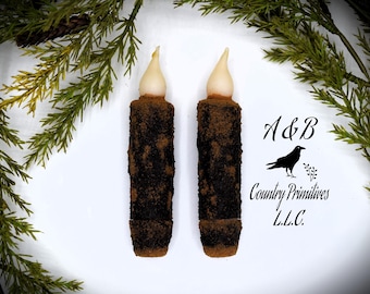 May include: Two brown and black pillar candles with white wax flames. The candles are on a white background with green foliage around the edges. The text "A&B Country Primitives LLC." is in the lower right corner of the image.