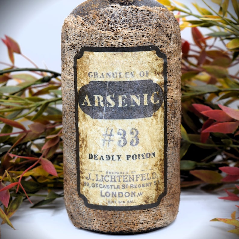 An antique brown bottle labeled "Granules of Arsenic #33 Deadly Poison" is in front of autumn-colored foliage. The bottle has a rustic appearance.