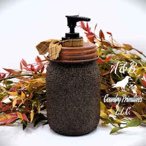 May include: A brown glass soap dispenser with a black pump and a rusty brown lid. The dispenser is decorated with a plaid ribbon and has a textured surface. The dispenser is sitting on a white surface with a branch of fall leaves behind it. The text "A&B Country Primitives L.L.C." is visible in the background.