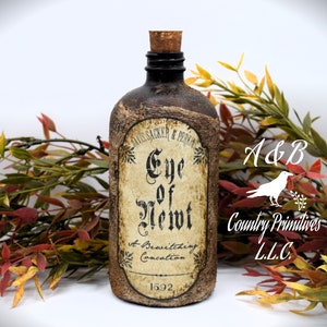 An antique brown bottle labeled "Eye of Newt, A Bewitching Collection" is in front of autumn-colored foliage. The bottle has a cork stopper and rustic appearance.