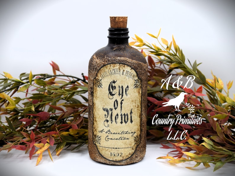An antique brown bottle labeled "Eye of Newt, A Bewitching Collection" is in front of autumn-colored foliage. The bottle has a cork stopper and rustic appearance.