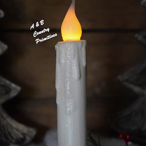 Zoomed in view of the top half of white glittered taper candle, focusing on glowing, flame shaped, wax dipped candle bulb, realistic looking wax drips down sides of candle, and shimmering glitter coating.  Winter decor in background.