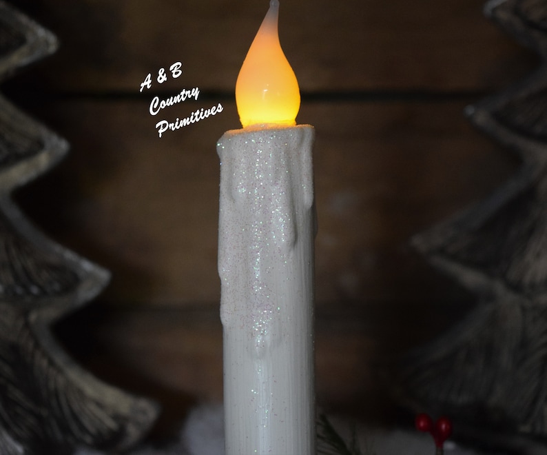 Zoomed in view of the top half of white glittered taper candle, focusing on glowing, flame shaped, wax dipped candle bulb, realistic looking wax drips down sides of candle, and shimmering glitter coating.  Winter decor in background.