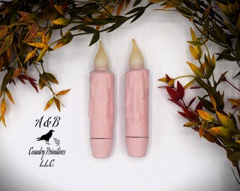 May include: Two pink taper candles with a textured, matte finish and a faux melted wax drip design. Each candle features a rounded, off-white flame-shaped tip at the top. The text A&B Country Primitives L.L.C. with a crow icon is visible.