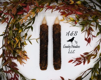 May include: Two brown and black taper candles with a speckled finish. The candles are lit and have a warm glow. The candles are surrounded by fall foliage. A&B Country Primitives L.L.C.
