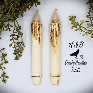 Puede incluir: Dos velas votivas blancas falsas con un efecto de goteo marrón y una llama de plástico transparente. Las velas están sobre un fondo blanco con follaje verde.