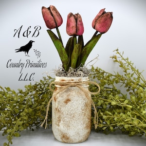 May include: A white distressed mason jar with a burlap bow and twine, filled with pink tulips and dried moss. The jar is sitting on a white surface with green foliage in the background. The text "A&B Country Primitives L.L.C." is visible in the image.