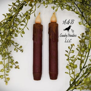 Puede incluir: Dos velas LED rojas con un brillo blanco cálido. Las velas miden aproximadamente 15 cm de alto y tienen un acabado rústico y desgastado. Las velas están rodeadas de follaje verde.