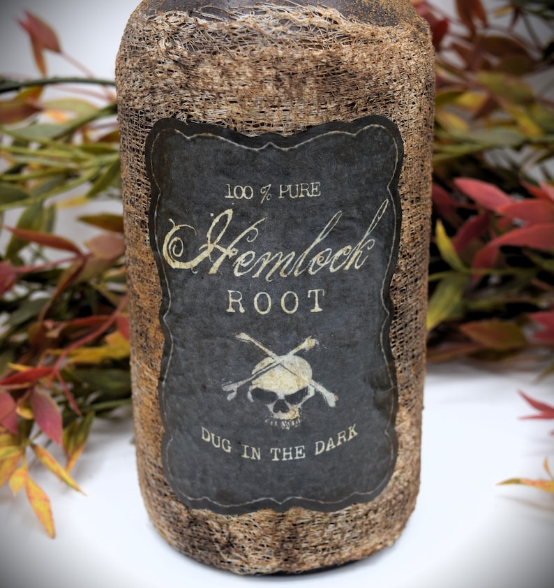 An antique brown bottle labeled "100% Pure Hemlock Root Dug in the Dark" with a skull and crossbones symbol is in front of autumn-colored foliage. The bottle has a rustic appearance.