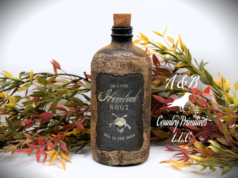 An antique brown bottle labeled "100% Pure Hemlock Root Dug in the Dark" with a skull and crossbones symbol is in front of autumn-colored foliage. The bottle has a cork stopper and rustic appearance.