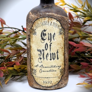 An antique brown bottle labeled "Eye of Newt, A Bewitching Collection" is in front of autumn-colored foliage. The bottle has a rustic appearance.