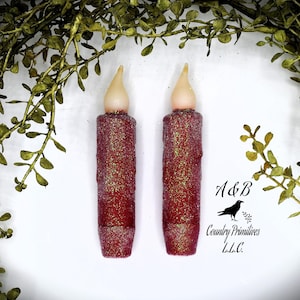 May include: Two red glitter battery-operated pillar candles with a warm white LED light. The candles are approximately 3 inches tall and have a gold glitter finish.