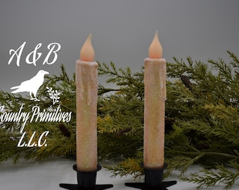 May include: Two white pillar candles with a glittery finish and a warm white LED flame. The candles are on black candle holders with a star-shaped base. The candles are surrounded by green foliage.