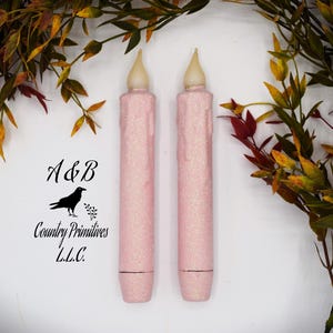 May include: Two cylindrical pink taper candles with a glittery finish and a faux melted wax texture. Each candle features a cream-colored flame-shaped bulb at the top and a flat base. The text A&B Country Primitives L.L.C. with a crow icon is visible.