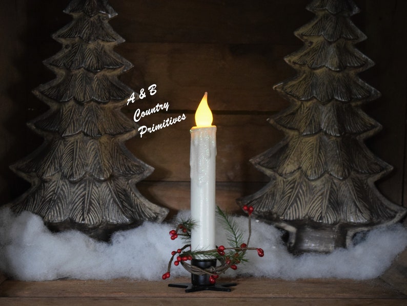 7 inch, white taper candle coated in shimmering glitter, with wax dipped, flame shaped candle bulb  glowing yellow/ orange, displayed in black wrought iron, star shaped holder, with decorative pine candle ring.  Fake snow and Christmas decor behind.