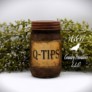 Könnte beinhalten: Ein braunes Glasgefäß mit einem rostigen braunen Deckel und einem Etikett mit der Aufschrift "Q-TIPS". Das Gefäß ist mit einem braunen, sackartigen Material verziert. Das Gefäß steht auf einer weißen Oberfläche. Der Text "A&B Country Primitives L.L.C." ist im Hintergrund sichtbar.