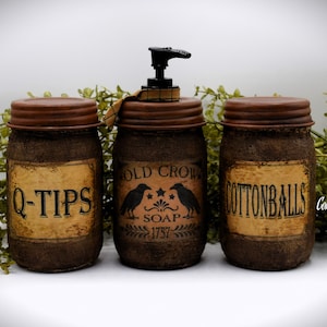 Op de afbeelding: Drie bruine glazen potten met jutehoezen en zwarte etiketten. De middelste pot is een zeepdispenser met een zwarte pomp en een etiket met de tekst "Old Crow Soap 1757". De pot links heeft een etiket met de tekst "Q-Tips" en de pot rechts heeft een etiket met de tekst "Cottonballs".