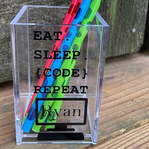 May include: A clear acrylic pencil holder with the text "Eat. Sleep. {Code} Repeat" and the name "Ryan" printed on it. The holder contains four pencils with colorful polka dot patterns.