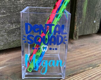 May include: A clear acrylic pencil holder with a blue and white "Dental Squad" design and a turquoise "Megan" name. The holder has three pencils inside, one green, one pink, and one blue, all with white polka dots.