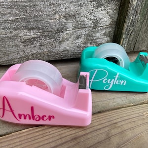 May include: Two personalized tape dispensers, one pink with the name "Amber" and one teal with the name "Peyton".