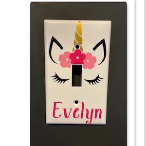 May include: White light switch cover with a unicorn design. The unicorn has a gold horn, pink flowers, and black eyelashes. The name "Evelyn" is written in pink script below the switch opening.