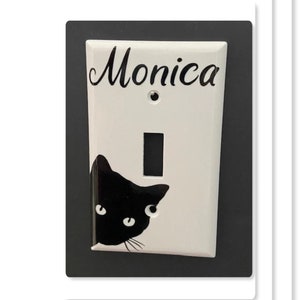 May include: White light switch cover with a black cat peeking out from the bottom and the name "Monica" written in cursive at the top.