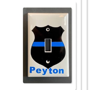 May include: White light switch cover with a black and blue police badge design. The badge has a thin blue line across the center. The name "Peyton" is printed in blue below the badge.