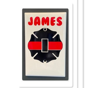 May include: White light switch cover with a black and red firefighter's thin red line emblem and the name "JAMES" in red letters.