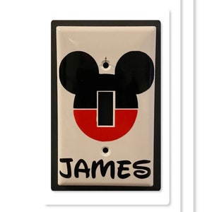 May include: A white light switch cover with a black Mickey Mouse head design and the name "JAMES" written in black below.