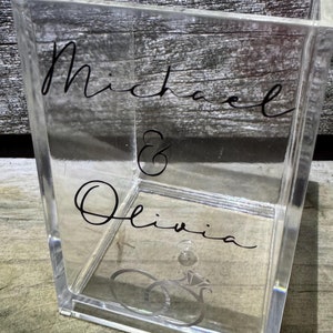 May include: A clear acrylic box with the names "Michael & Olivia" written in black cursive script. Two silver rings are inside the box.