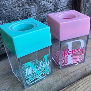 K&ouml;nnte beinhalten: Zwei durchsichtige Plastikbeh&auml;lter mit einem rosa und t&uuml;rkisfarbenen Deckel. Die Beh&auml;lter sind mit B&uuml;roklammern gef&uuml;llt und haben einen personalisierten Buchstaben und Namen auf der Vorderseite. Der t&uuml;rkisfarbene Beh&auml;lter sagt "Marley" und der rosa Beh&auml;lter sagt "Penny".