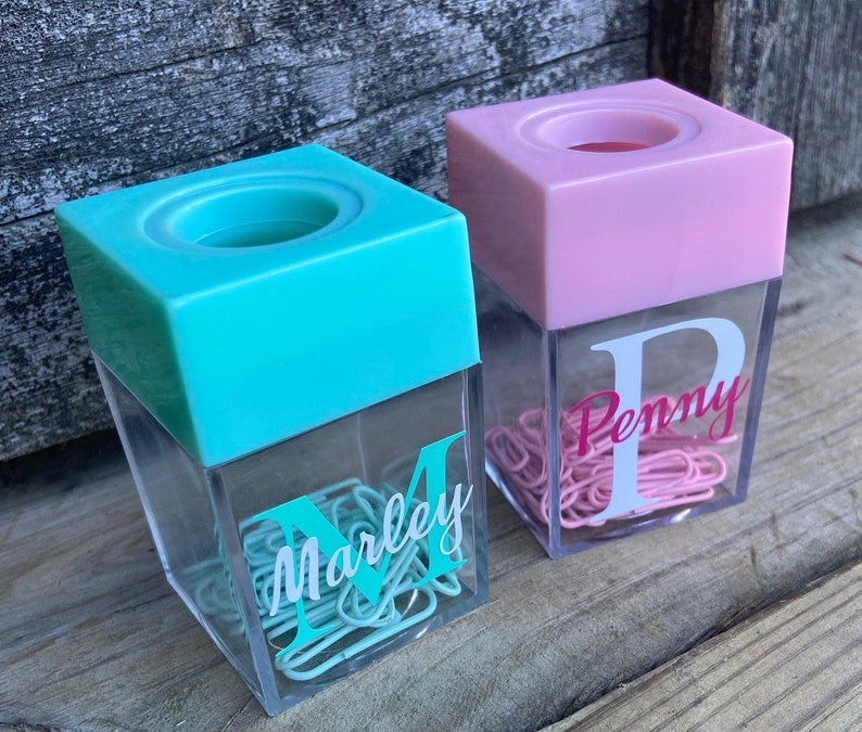 K&ouml;nnte beinhalten: Zwei durchsichtige Plastikbeh&auml;lter mit einem rosa und t&uuml;rkisfarbenen Deckel. Die Beh&auml;lter sind mit B&uuml;roklammern gef&uuml;llt und haben einen personalisierten Buchstaben und Namen auf der Vorderseite. Der t&uuml;rkisfarbene Beh&auml;lter sagt "Marley" und der rosa Beh&auml;lter sagt "Penny".