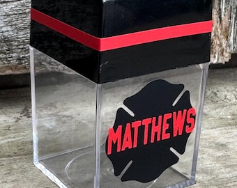 May include: Clear acrylic tissue box with a black top and red stripe. A black and red firefighter Maltese cross with the name "MATTHEWS" is on the front. The box has a square shape.