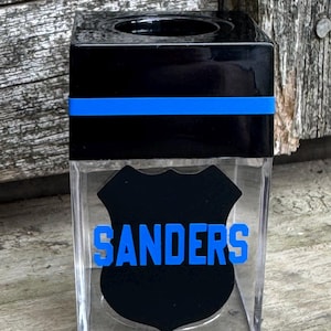 May include: A clear, square tissue box with a black top and a blue stripe. The front features a black shield with the name "SANDERS" in blue lettering. The design is likely related to law enforcement.