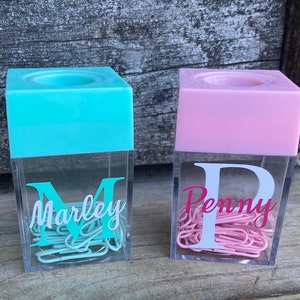 K&ouml;nnte beinhalten: Zwei durchsichtige Plastikbeh&auml;lter mit magnetischen Deckeln, einer t&uuml;rkisfarben und einer rosa, gef&uuml;llt mit B&uuml;roklammern. Die Beh&auml;lter haben einen wei&szlig;en Buchstaben und Namen auf der Vorderseite, "M Marley" und "P Penny".