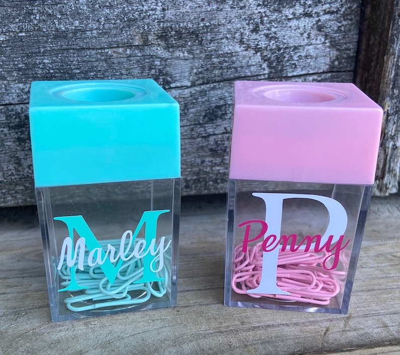 K&ouml;nnte beinhalten: Zwei durchsichtige Plastikbeh&auml;lter mit magnetischen Deckeln, einer t&uuml;rkisfarben und einer rosa, gef&uuml;llt mit B&uuml;roklammern. Die Beh&auml;lter haben einen wei&szlig;en Buchstaben und Namen auf der Vorderseite, "M Marley" und "P Penny".