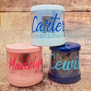 May include: Three personalized pencil sharpeners in pink, blue, and navy blue. Each sharpener has a clear plastic body and a colored lid and base. The names "Madelyn", "Carter", and "Lewis" are written on the front in cursive.