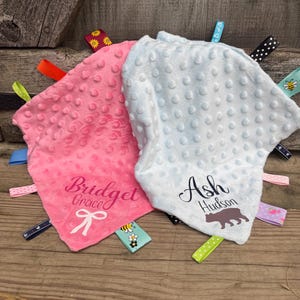 May include: Two baby security blankets, one pink and one light blue, with textured surfaces and colorful ribbon tags. The pink blanket has the name "Bridget Grace" and a bow graphic. The blue blanket has the name "Ash Hudson" and a bear graphic.