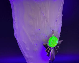 May include: A silver necklace with a green glass pendant. The pendant is a circle with a swirling pattern inside. The necklace is hanging from a white vase.