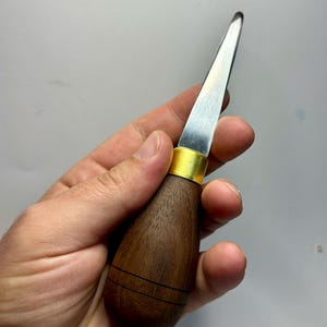 Puede incluir: Un primer plano de una pequeña herramienta de metal afilada con un mango de madera. El mango es de color marrón oscuro con una banda de latón en la parte superior. La herramienta se sostiene en una mano.