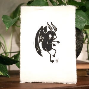 May include: Black and white illustration of a winged creature with large eyes, printed on a white card.