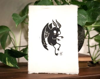 May include: Black and white illustration of a winged creature with large eyes, printed on a white card.