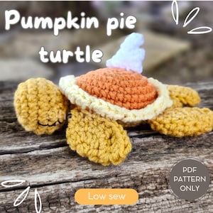 以下が含まれることがあります： 白いホイップクリームのトッピングをした、かぎ針編みのかぼちゃパイの亀のぬいぐるみです。亀は木製の表面に横たわっています。亀の下のボタンには「Low sew」という文字が書かれています。画像の右下隅には白い枠の中に「PDF PATTERN ONLY」という文字が書かれています。