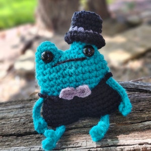 May include: A teal crocheted frog wearing a black top hat with a gray band and a gray bow tie. The frog is sitting on a wooden surface.